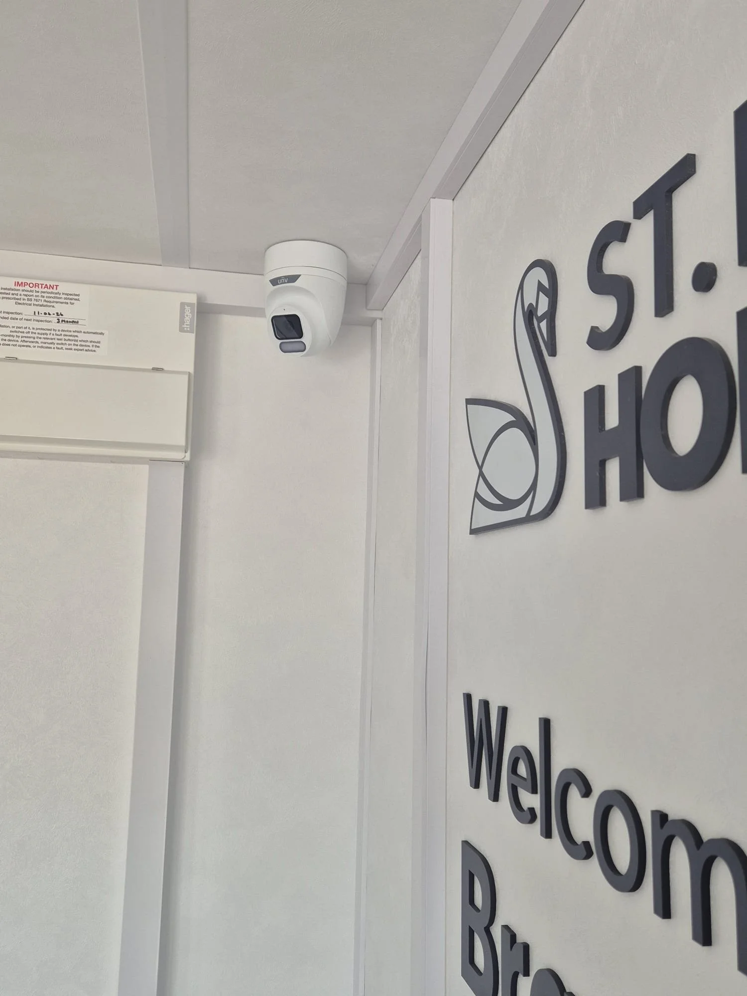 Security camera mounted on a ceiling in an office or lobby space. Partial wall sign with the word 'Welcome' and a stylized floral or decorative symbol next to it.
