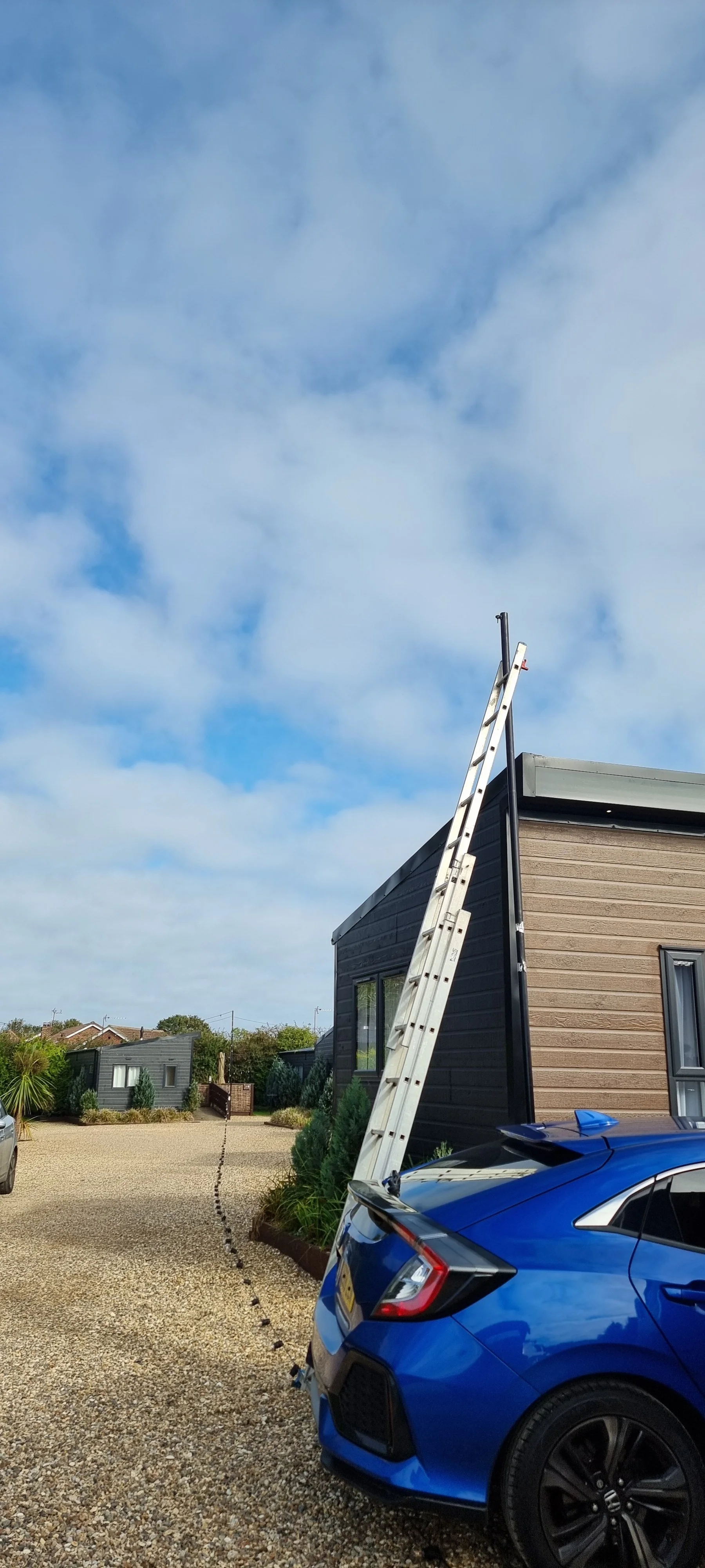 A white ladder leaning against the corner of a modern beige and black house with a small window. A blue car is parked in front of the house. The sky is partly cloudy.