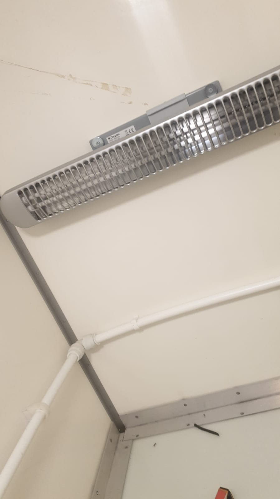Ceiling-mounted electric heating unit in a room corner with visible wiring and pipes.