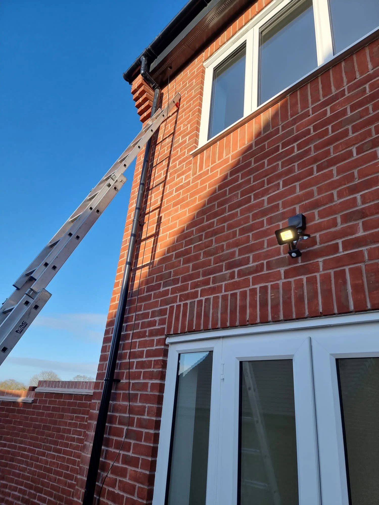 A ladder leaning against a brick house exterior under a clear blue sky, with a window, a black gutter, and a mounted outdoor light.