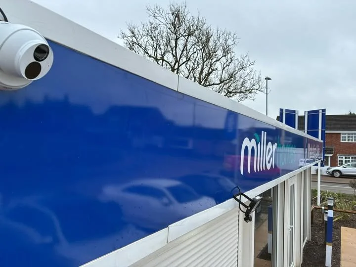 Exterior of a commercial building with a blue sign that reads "Miller" and a security camera mounted on the corner, with a tree and parked cars in the background.