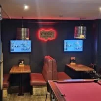 A dimly lit bar or lounge with a dark wall, two flat-screen TVs displaying sports, a neon Coca-Cola sign, and a pink pool table in the foreground. There are also two small hanging pendant lights and a booth-style seat.