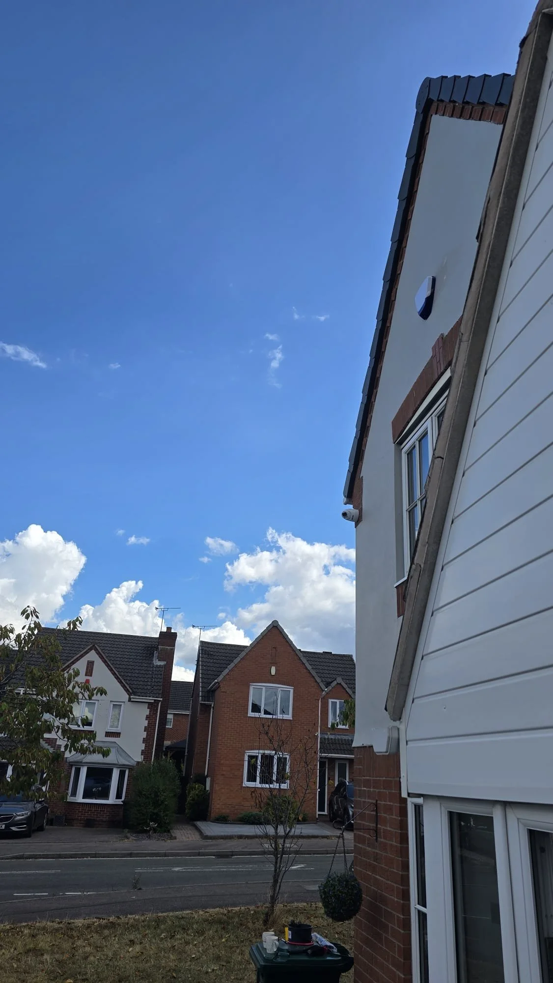 View of a residential neighborhood with red brick houses, trees, and a clear blue sky with some clouds, taken from a garden area next to a house.