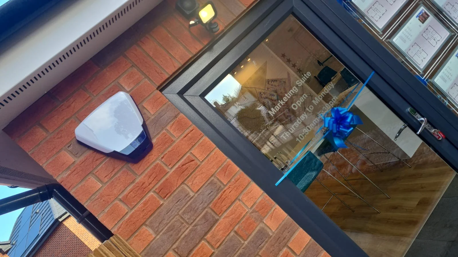 Outside view of a business with a brick wall, glass door with a blue ribbon decoration, and a sign indicating opening hours, with a white automatic hand sanitizer dispenser mounted on the wall nearby.