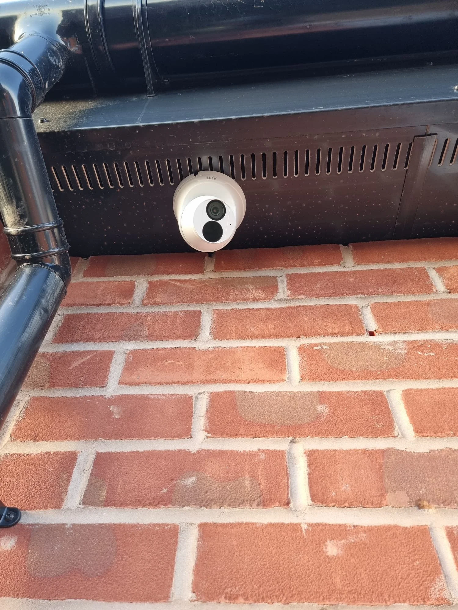 A white security or surveillance camera mounted underneath a black metal structure on a brick wall.