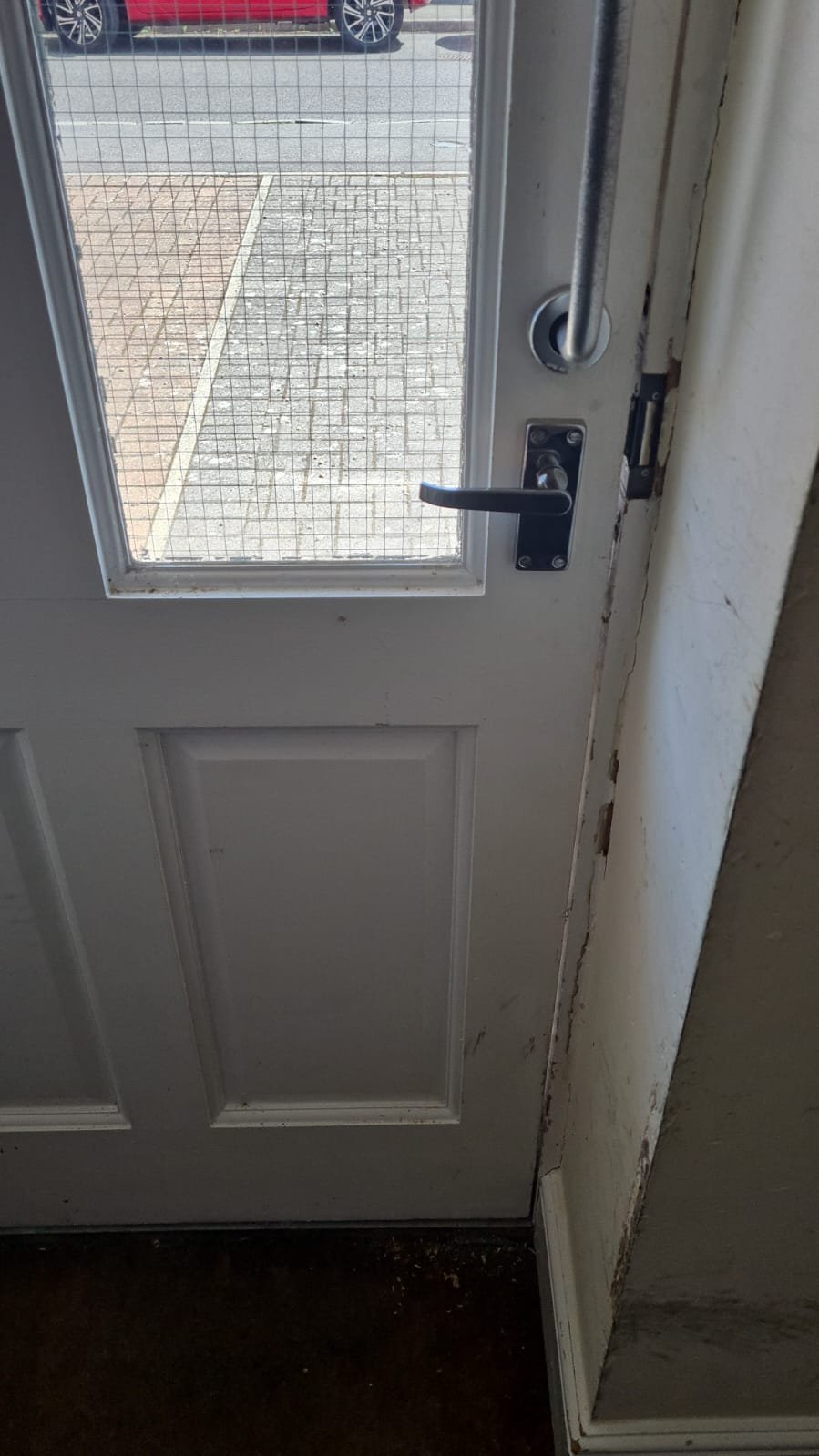 Close-up of a white door with a glass window and a black handle, leading outside to a parking lot with a red vehicle parked nearby.