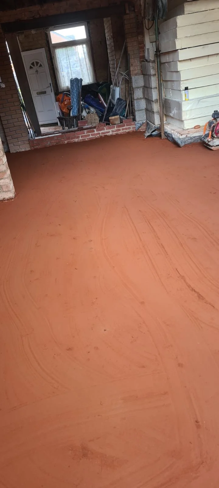 Interior of a house under construction with a newly leveled burnt orange floor, exposed brick walls, and construction tools and materials near a front door and window.