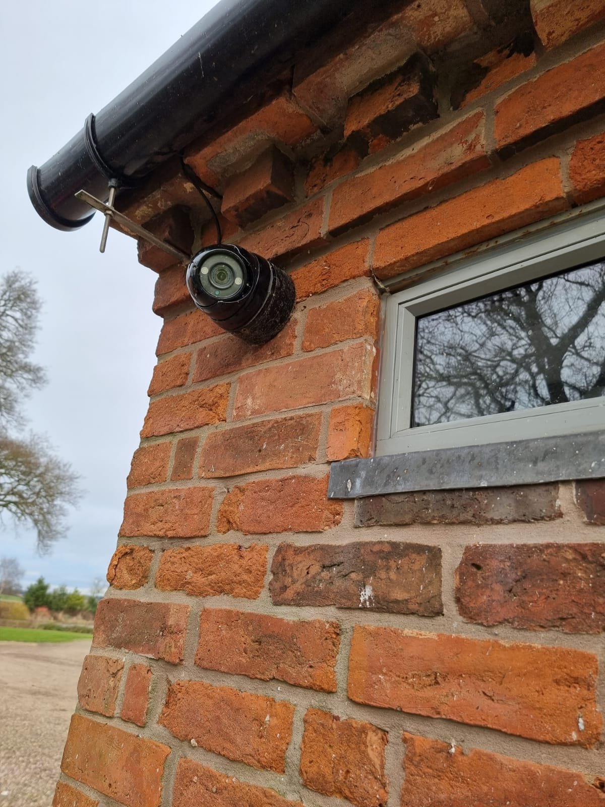 Close-up of a brick building corner with a small window, a black pipe, and a security camera mounted under the roof eaves, with trees and cloudy sky in the background.