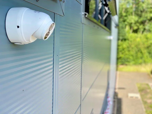 Close-up of a white security camera mounted on the side of a light blue building with horizontal ridges, with greenery and pavement in the background.