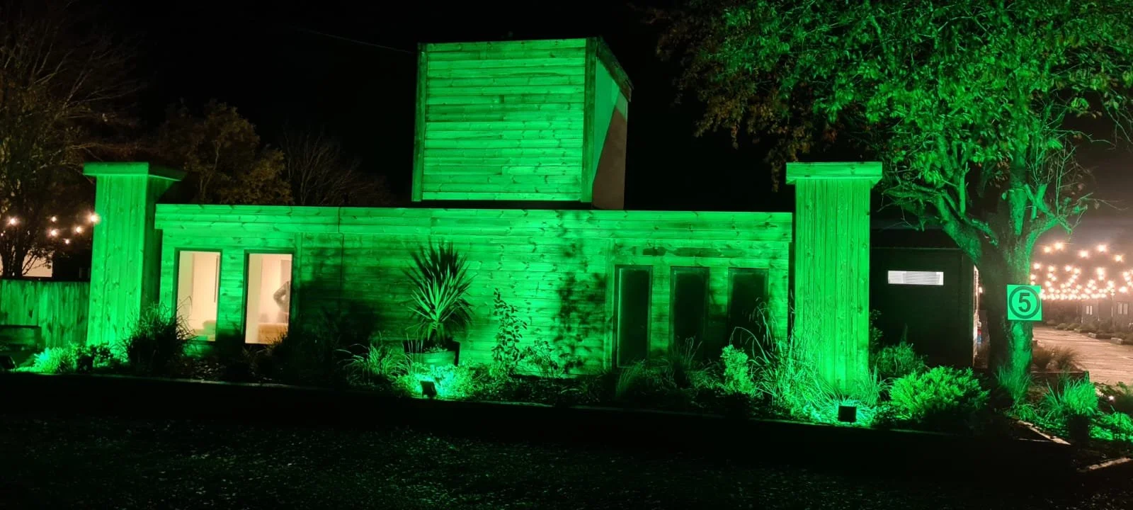 A house decorated with vibrant green exterior lights at night, with surrounding plants and trees illuminated, and a sidewalk with a numbered marker.
