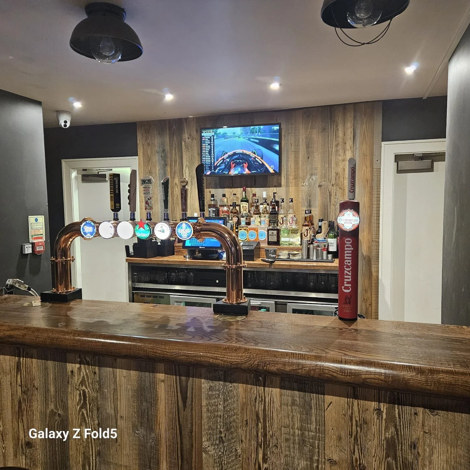 A bar with a wooden counter, beer taps, and a television screen displaying a racing game. There are bottles of liquor behind the bar and some bar tools. The wall behind the bar is also wood, and there's a tall Cruzcampo beer tower on the right.