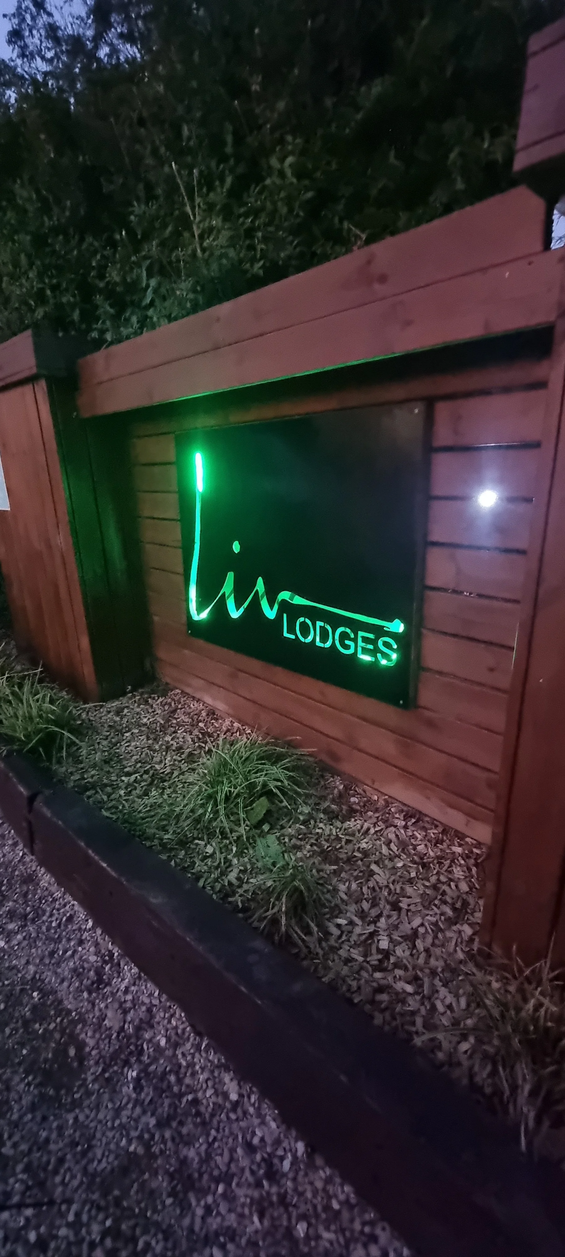 Neon sign for Liw Lodges displayed on a wooden wall with greenery at night.