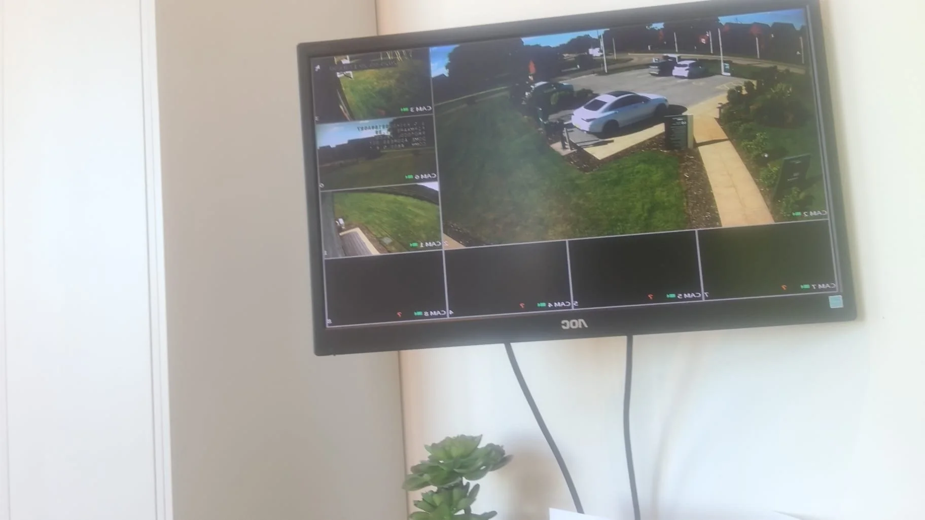 Security monitor displaying multiple camera feeds of a parking lot with cars and surrounding greenery.