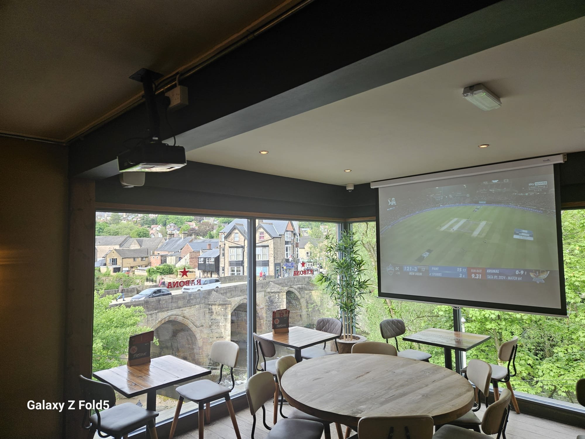 Interior of a dining area with several tables and chairs, large windows showing a street scene with houses and a bridge outside, and a large screen displaying a cricket match.