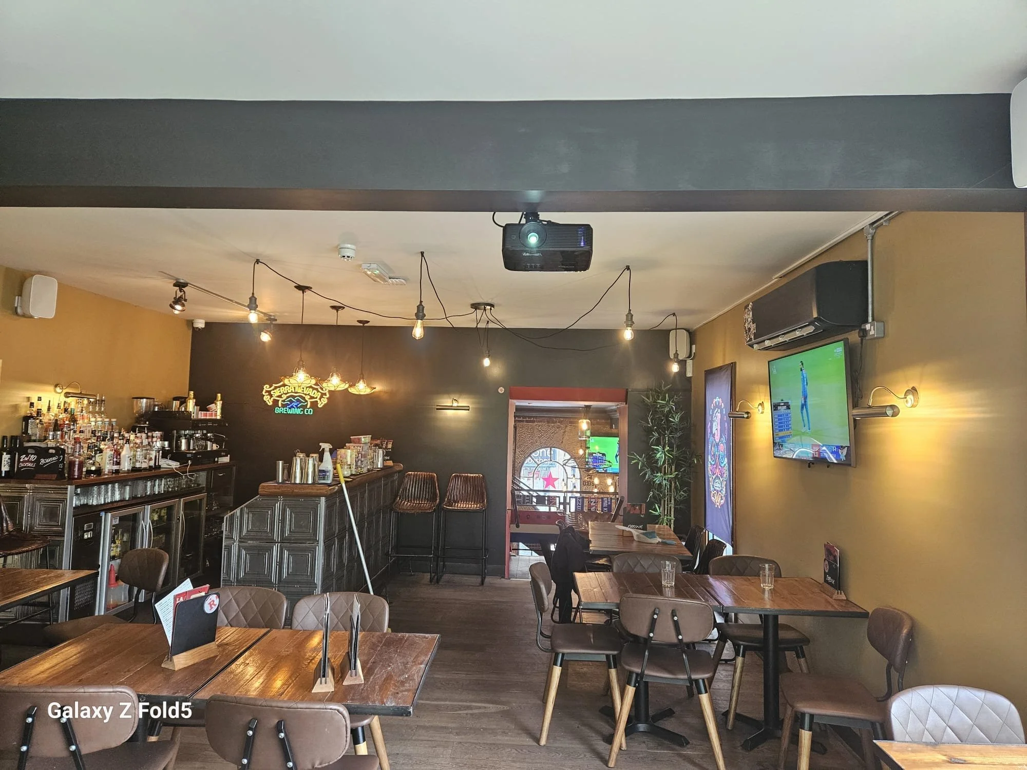 The image depicts the interior of a cozy cafe or restaurant with wooden tables and chairs, a bar area with liquor bottles, decorative hanging lights, a television on the wall, and some potted plants. The ceiling has a projector mounted, and the walls