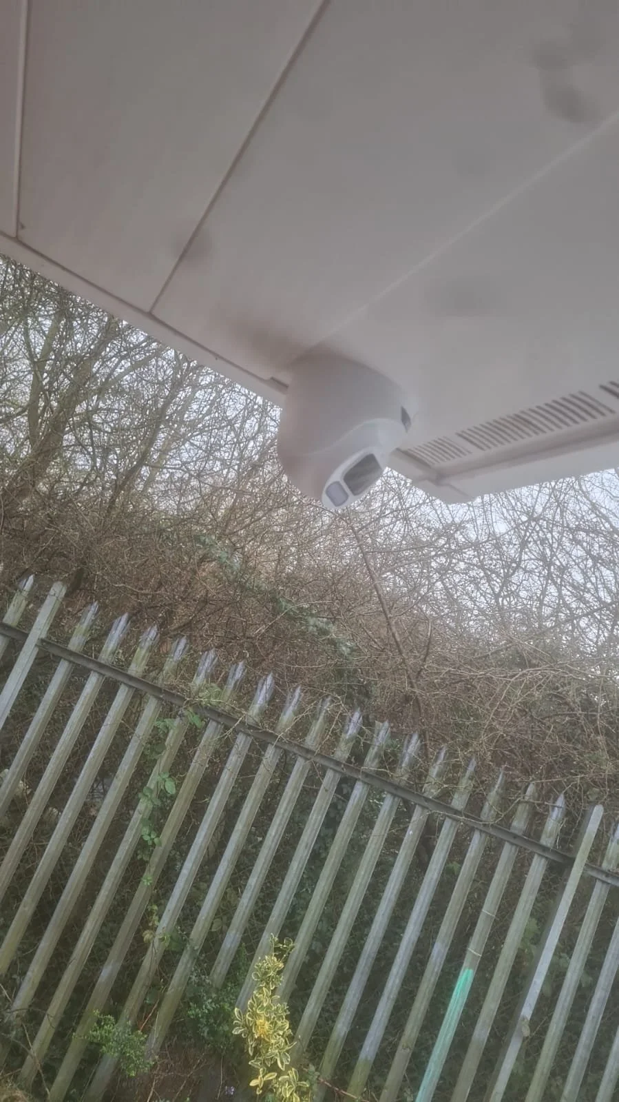 Security camera mounted under the edge of a white building roof, with a background of a brown wooden fence and leafless trees.