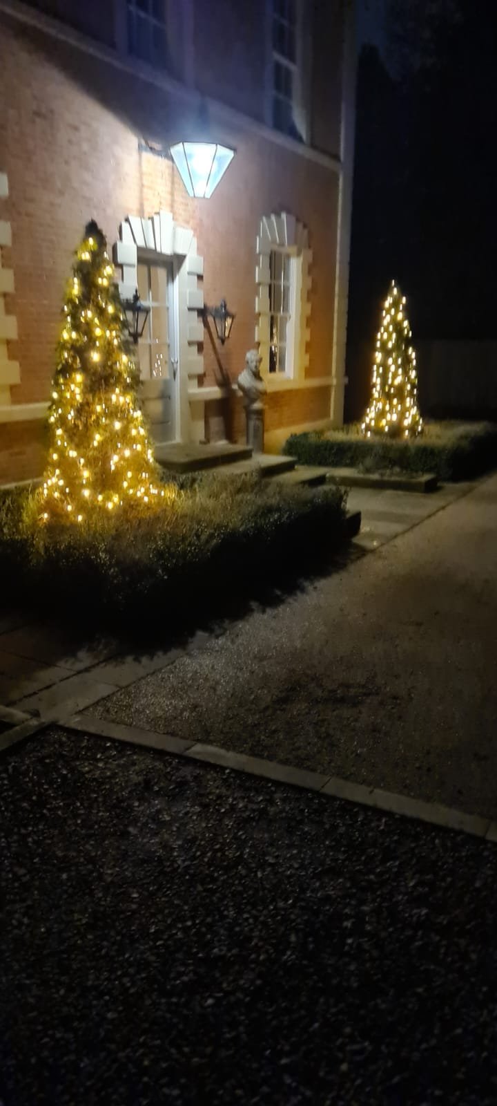 Nighttime scene showing two lit Christmas trees outside a brick house with two windows, a streetlamp, and a statue on the porch.