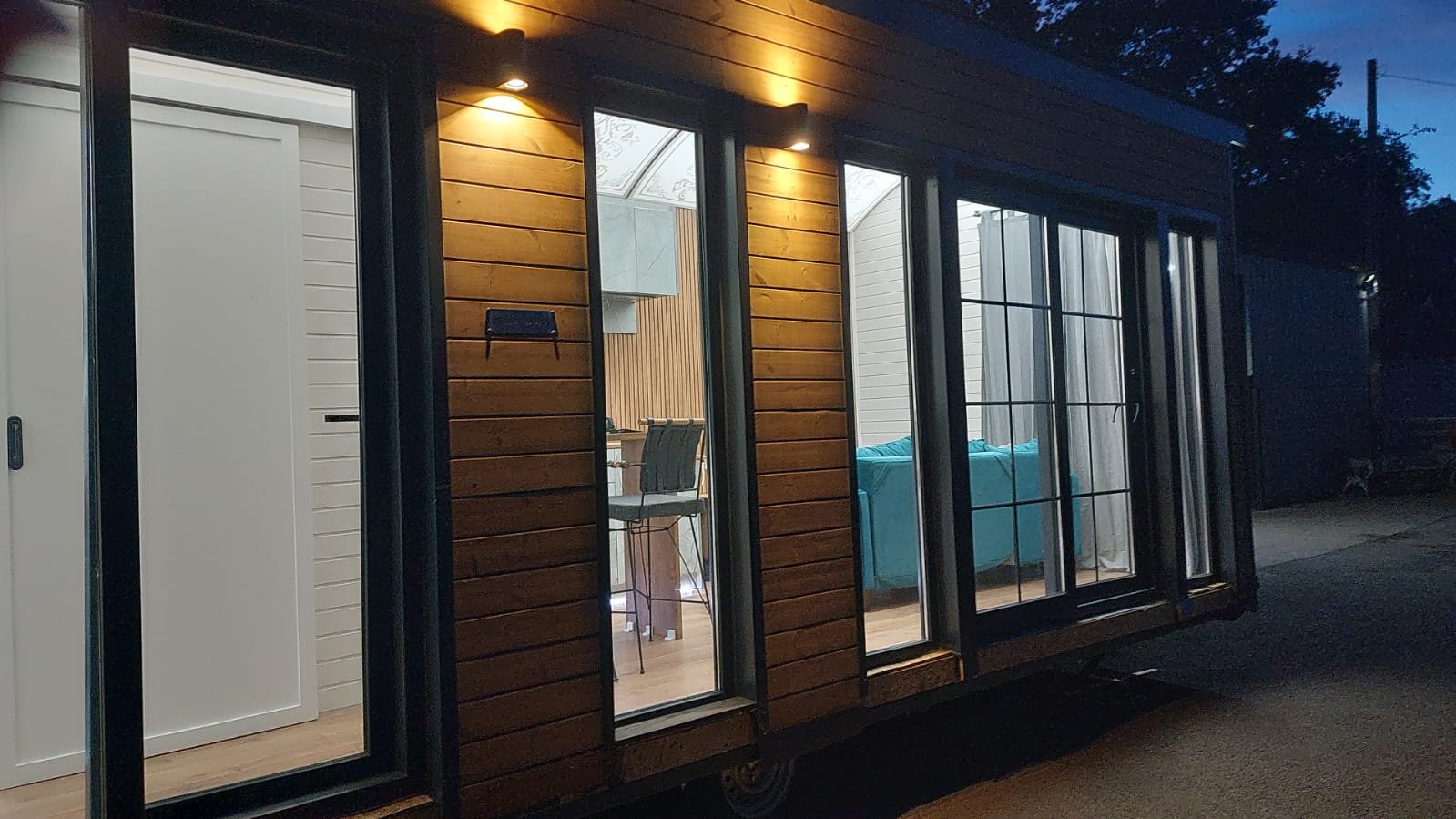 A tiny house on wheels with sliding glass doors, interior living space with a blue sofa, dining table, chairs, and a cloudy sky outside during dusk.