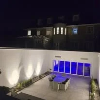 Nighttime view of a modern backyard patio with blue lighting, outdoor dining table with chairs, and a glass door leading inside.