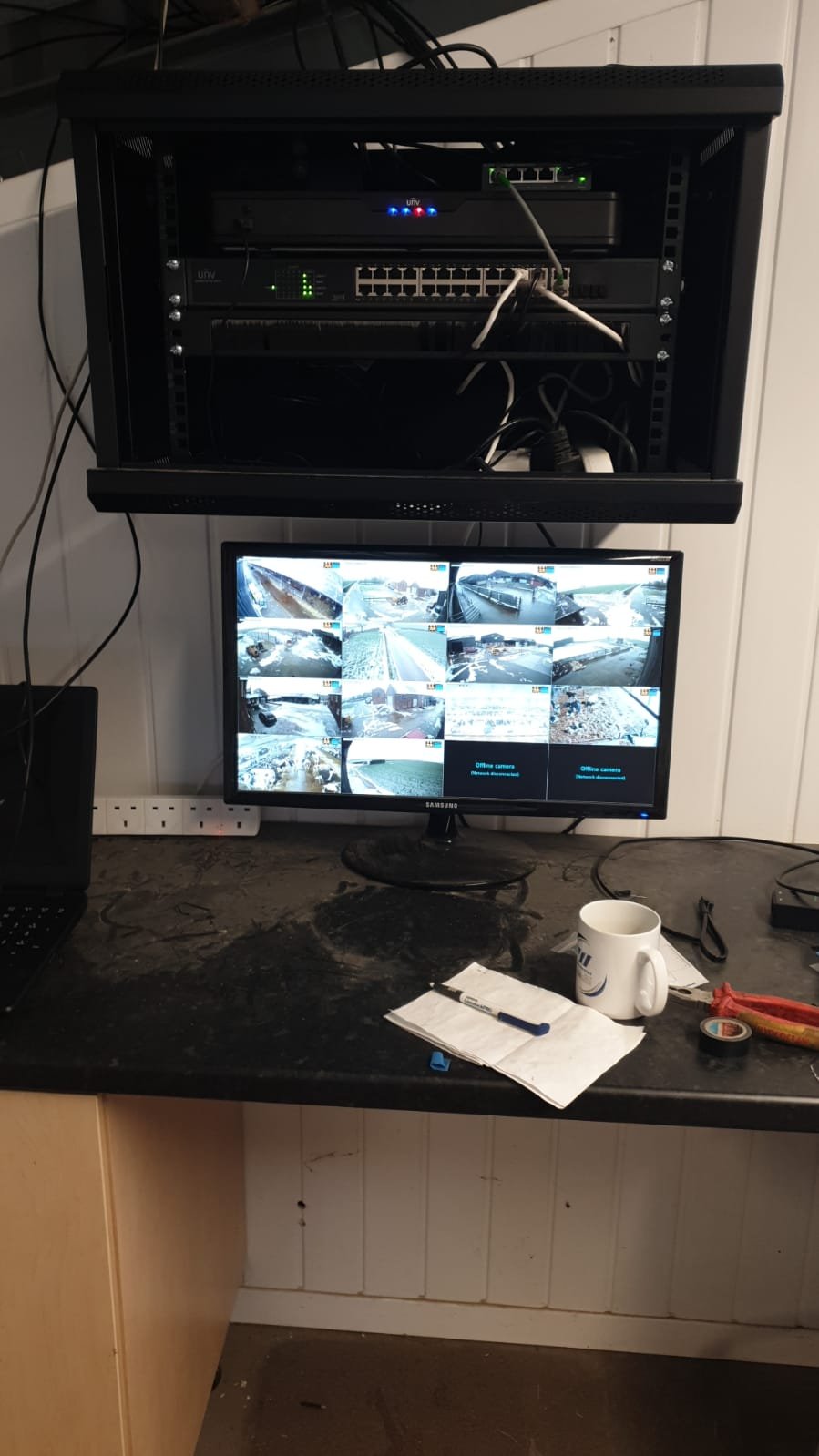 A surveillance monitor displaying multiple outdoor camera feeds, with a computer server rack mounted above it, on a cluttered black work surface with a mug, papers, pen, and tools.