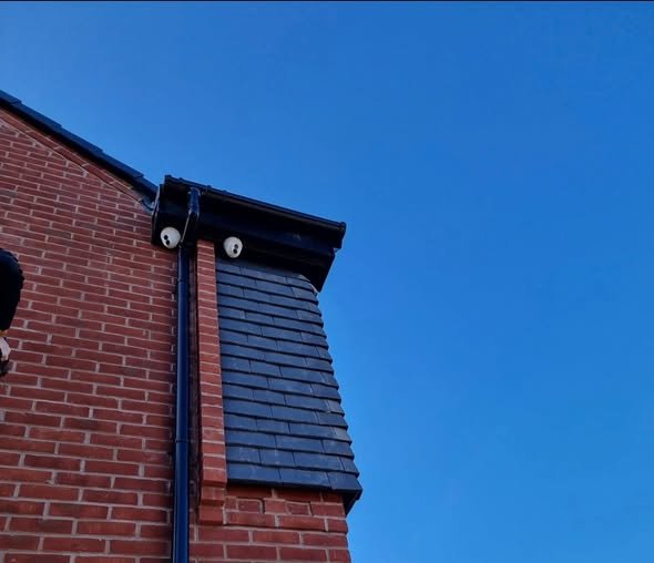 Close-up of the corner of a brick building with a blue slate roof, two security cameras, and a clear blue sky in the background.