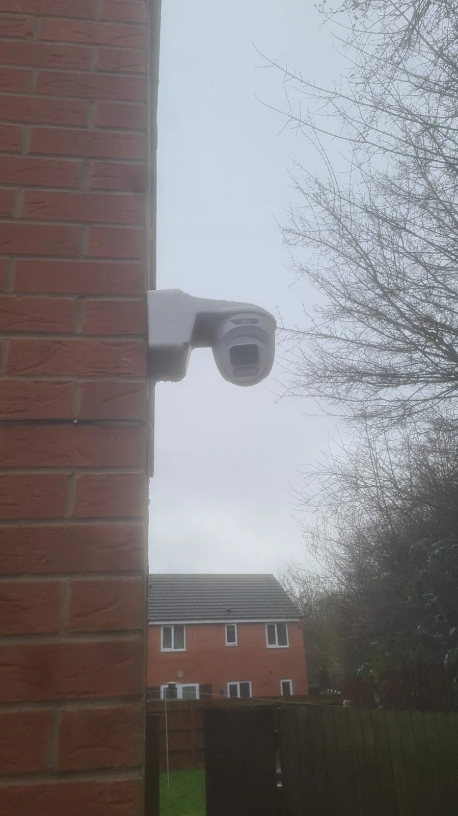 Security camera mounted on a brick wall, with houses and leafless trees in the background under an overcast sky.