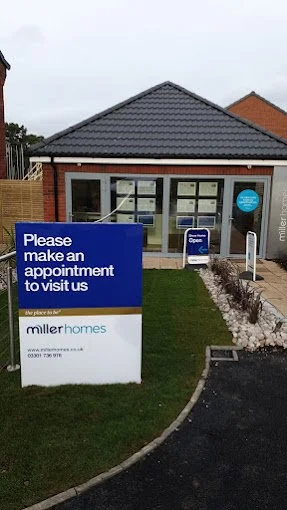 A construction or real estate office building with large windows, with a blue sign in front that reads, 'Please make an appointment to visit us.' There is also a smaller sign that says 'We are open.' The building is surrounded by a small garden with 