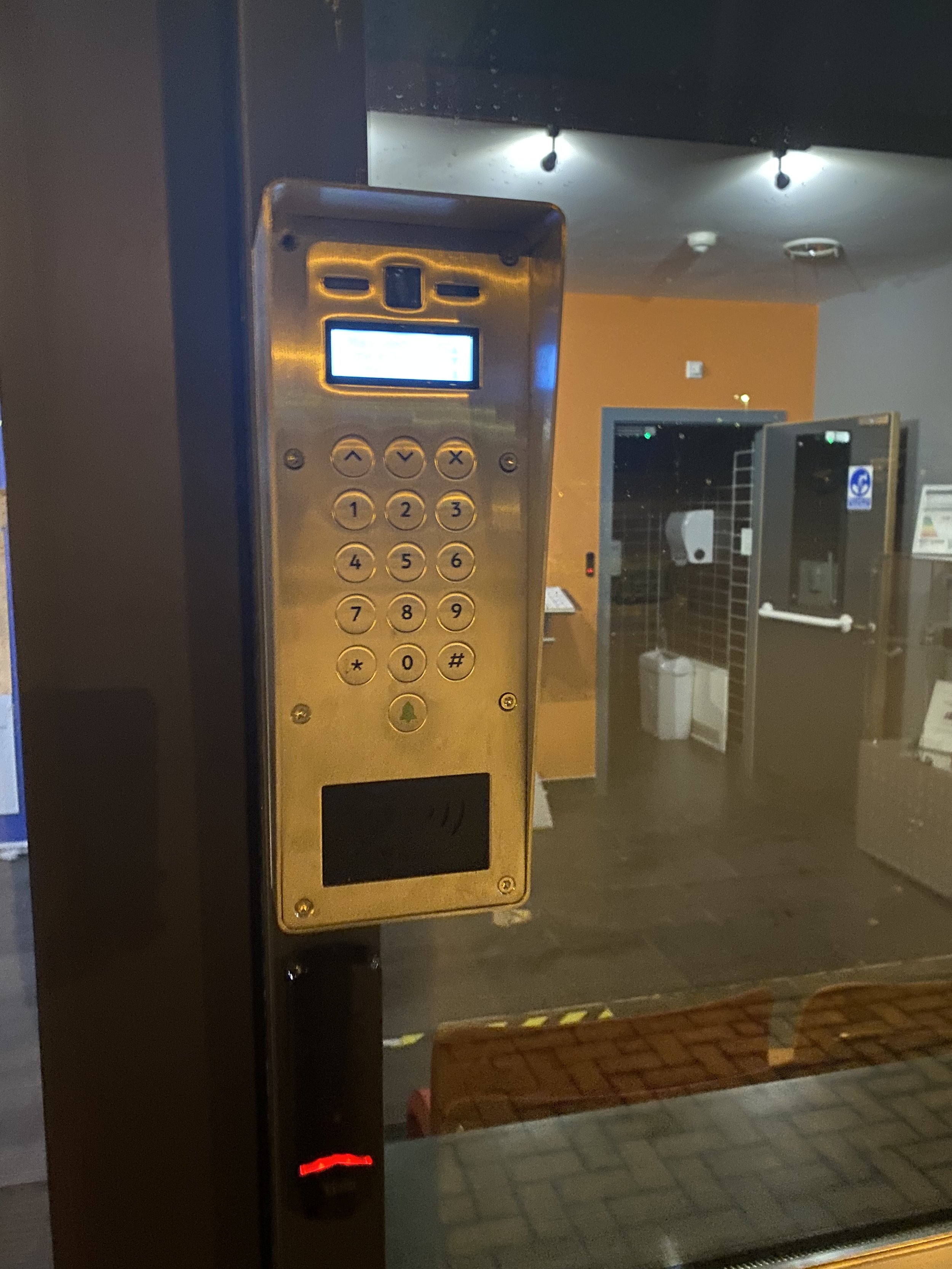 Electronic intercom or access control panel with a fingerprint scanner, keypad, and small display screen, mounted on a black pillar outside a building.