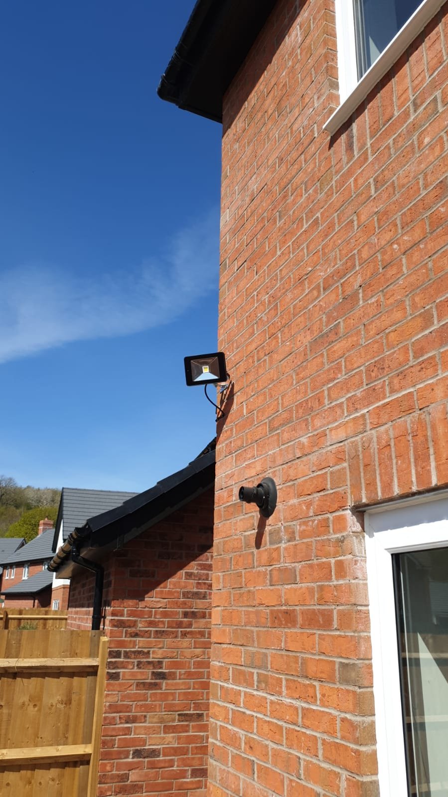 Exterior view of a brick house with outdoor security lighting, a window with a white frame, a gutter, and a neighboring house with similar design under a clear blue sky.