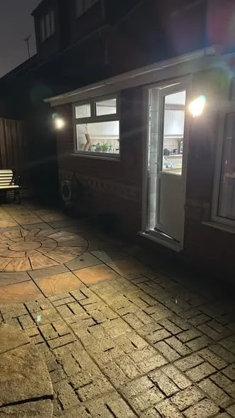 Backyard patio area with stone pavers, an exterior door, a window, and a bench, illuminated by outdoor lighting, during evening or night.