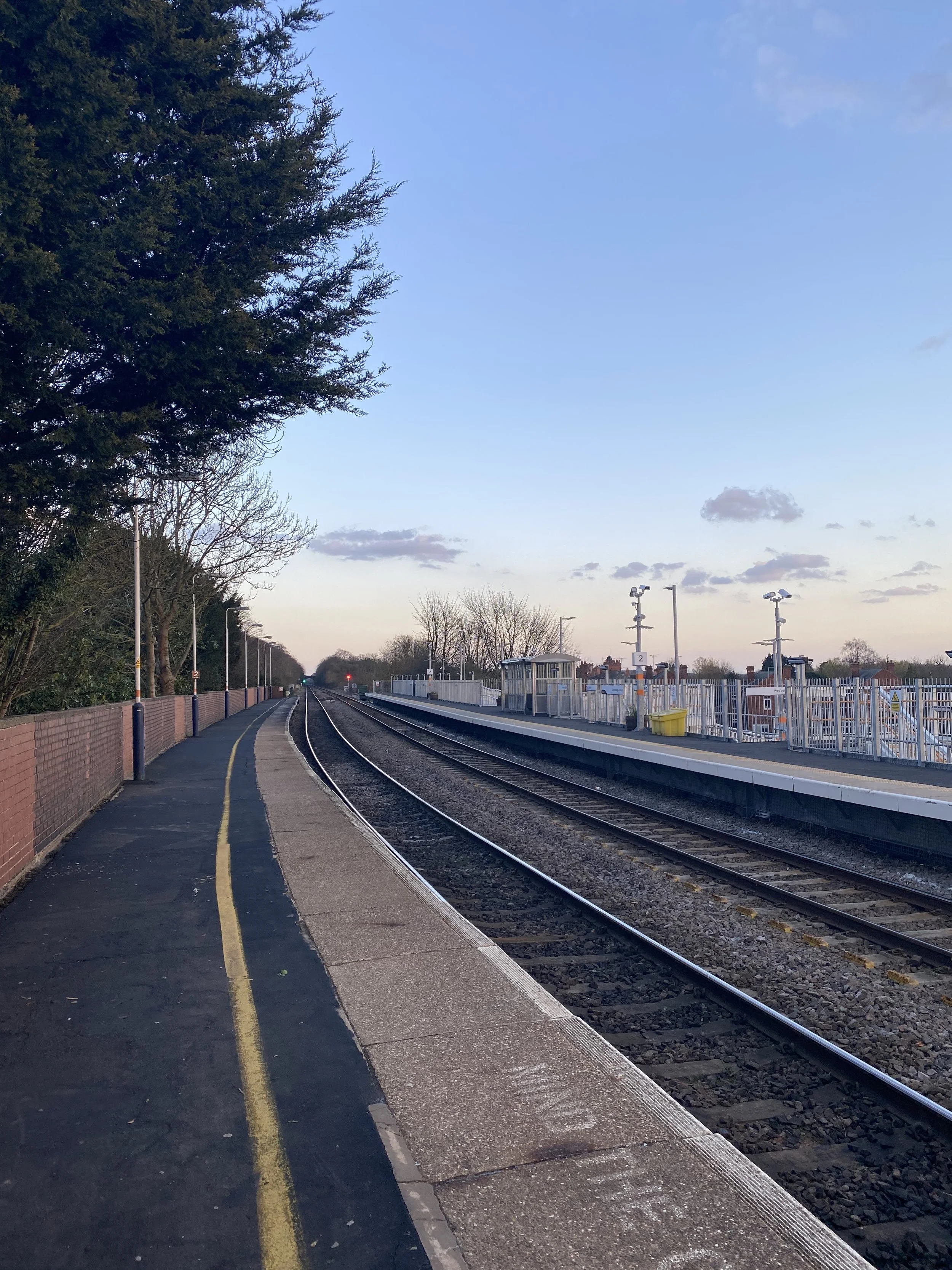A railway platform with tracks on the right, a paved walkway on the left bordered by a brick wall, and trees in the background. There are several street lamps and a small shelter on the platform. The sky is clear with a few clouds, indicating late af