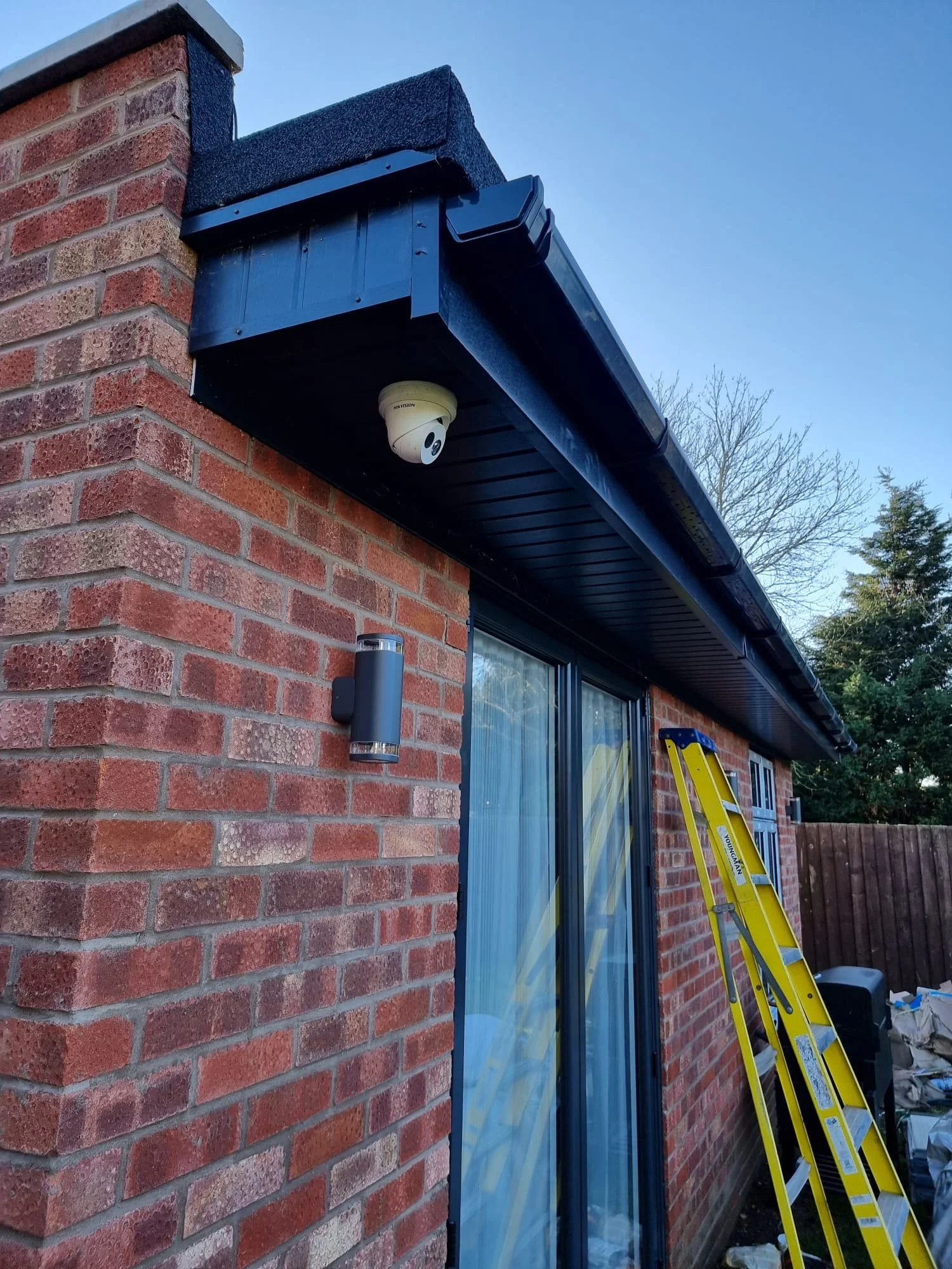 The exterior corner of a house with a brick wall and black trim. A security camera is mounted under the eaves, and a yellow ladder is leaning against the wall. There is a small outdoor light fixture nearby, and a window with a reflection and curtain.