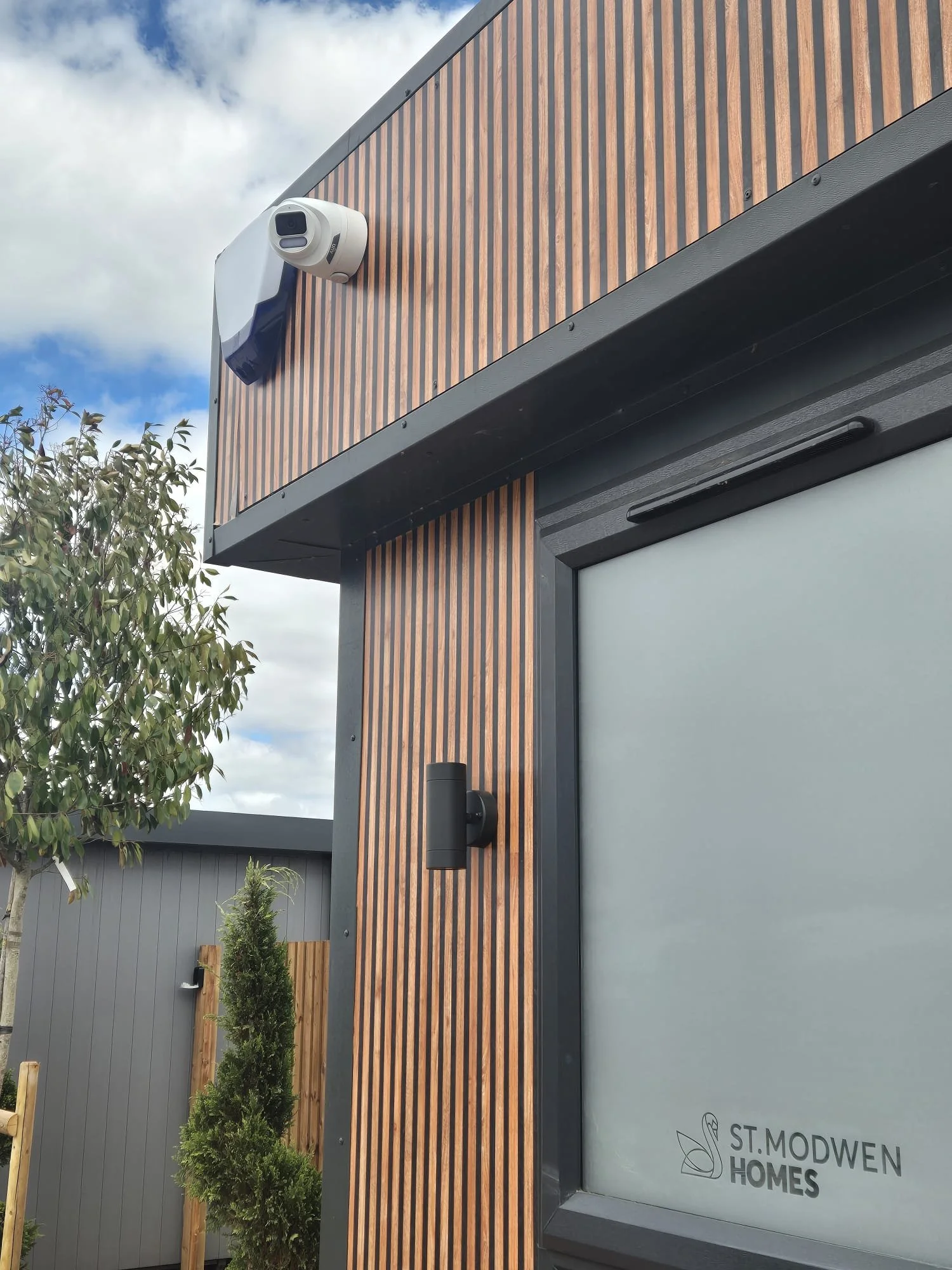 Modern house exterior with wooden siding, a security camera, a black outdoor light, and a window with the logo and name 'St. Modwen Homes'.