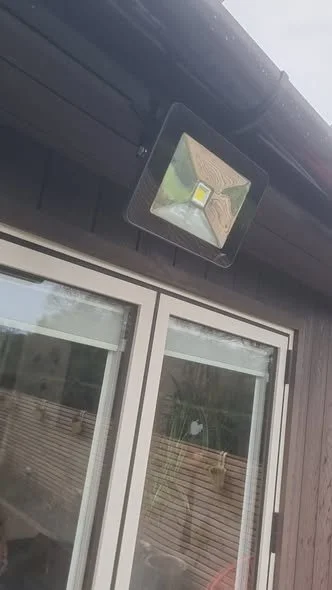 Outdoor security or floodlight mounted above a sliding glass door on a dark-colored building exterior.