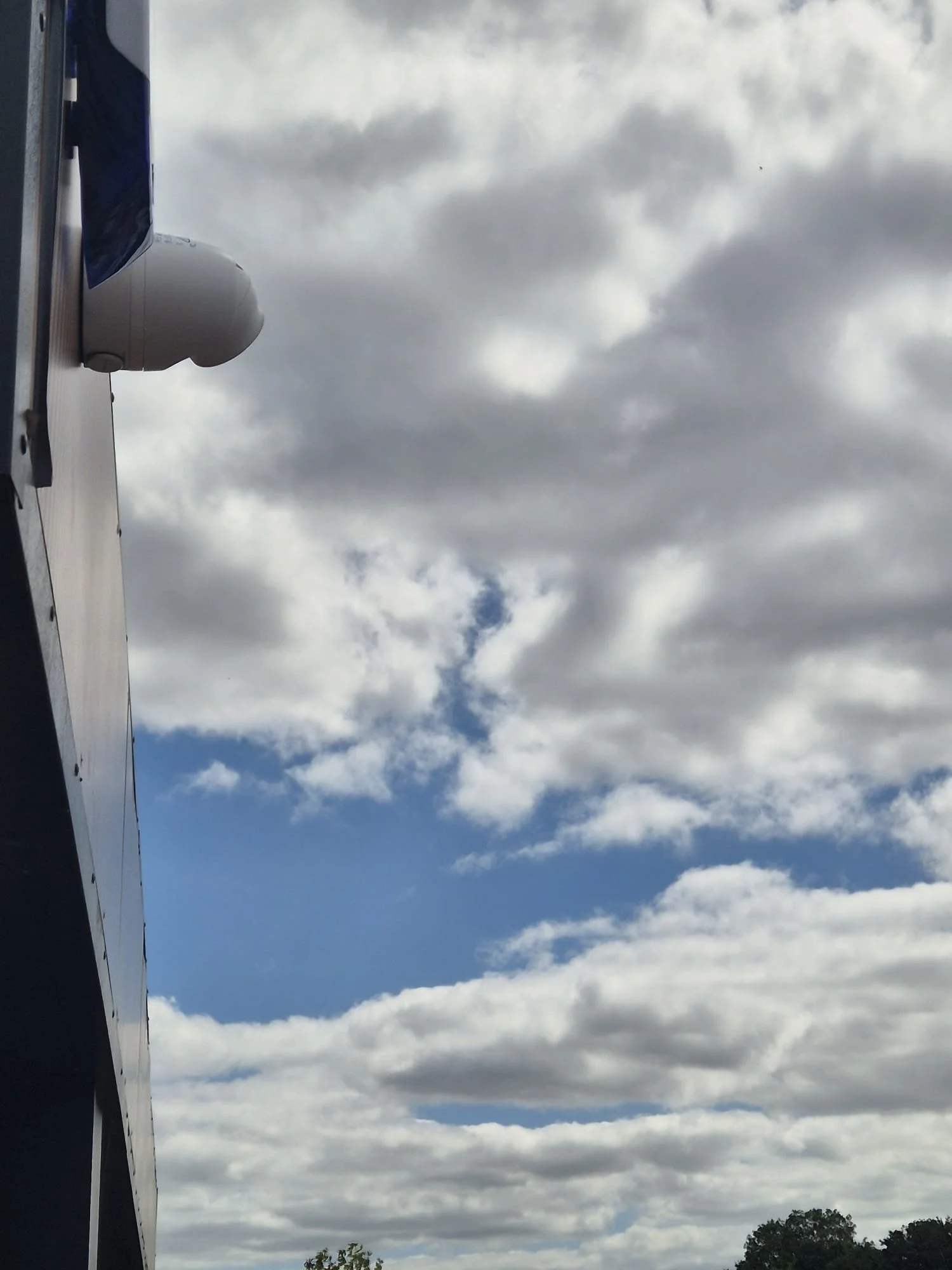 Part of an airplane wing with a weather radar, flying under a cloudy sky with some small trees visible at the bottom.