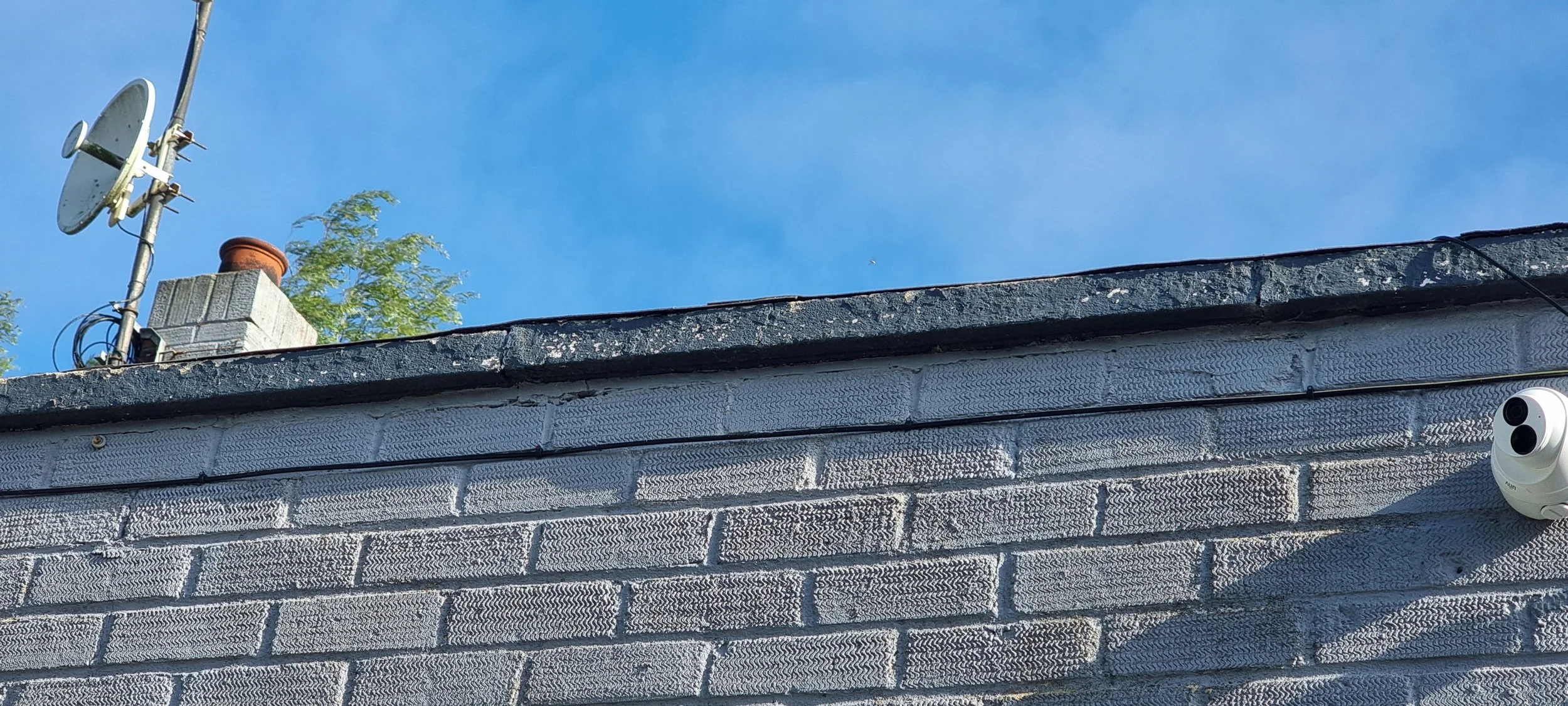 The rooftop of a building with a brick chimney, satellite dish, wires, and a security camera against a blue sky with a small tree in the background.