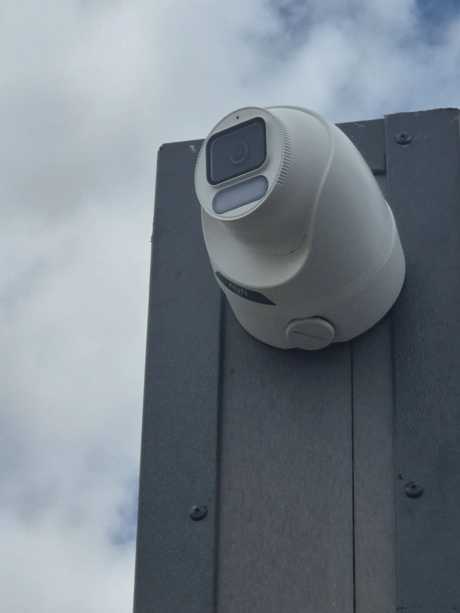 Close-up of a weatherproof security camera mounted on the corner of a building, with gray siding and a cloudy sky in the background.