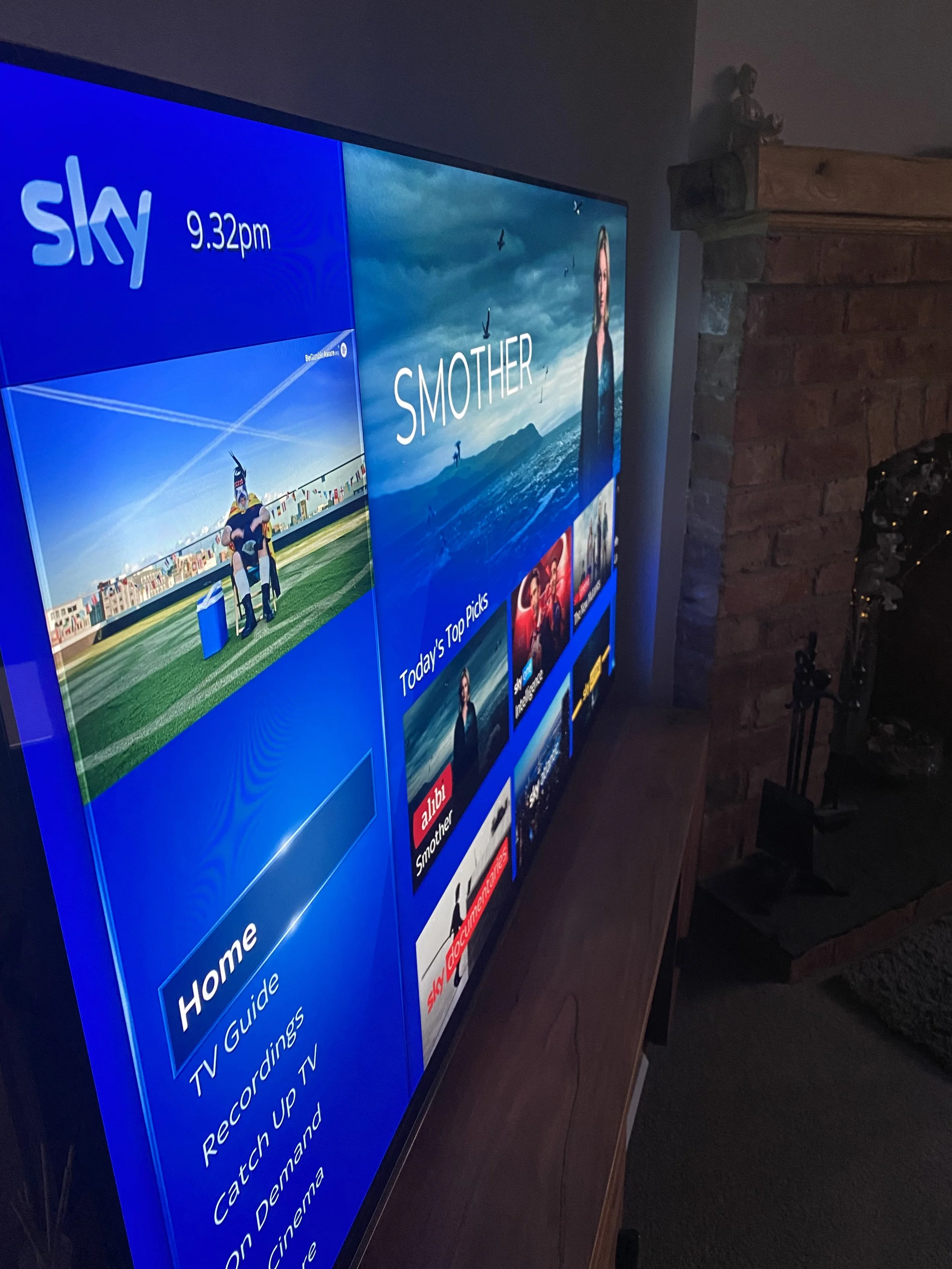 A digital TV or monitor displaying the Sky TV guide, showing current time as 9:32 pm, with various TV show options and a background image of a cloudy sky with birds, a mountain landscape, and a woman.