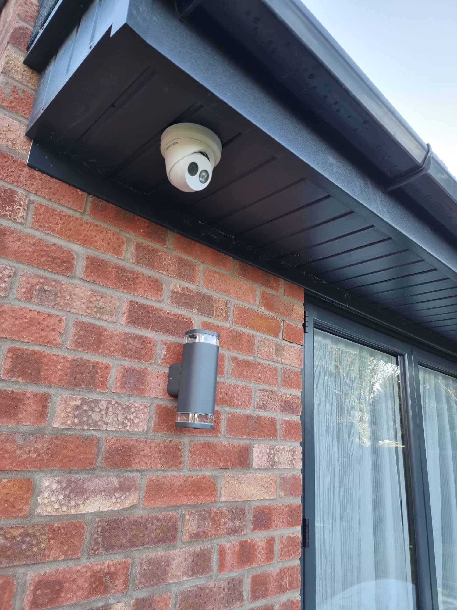 A security camera mounted under the eaves of a house, next to a wall-mounted outdoor light fixture, at the corner of a brick house.