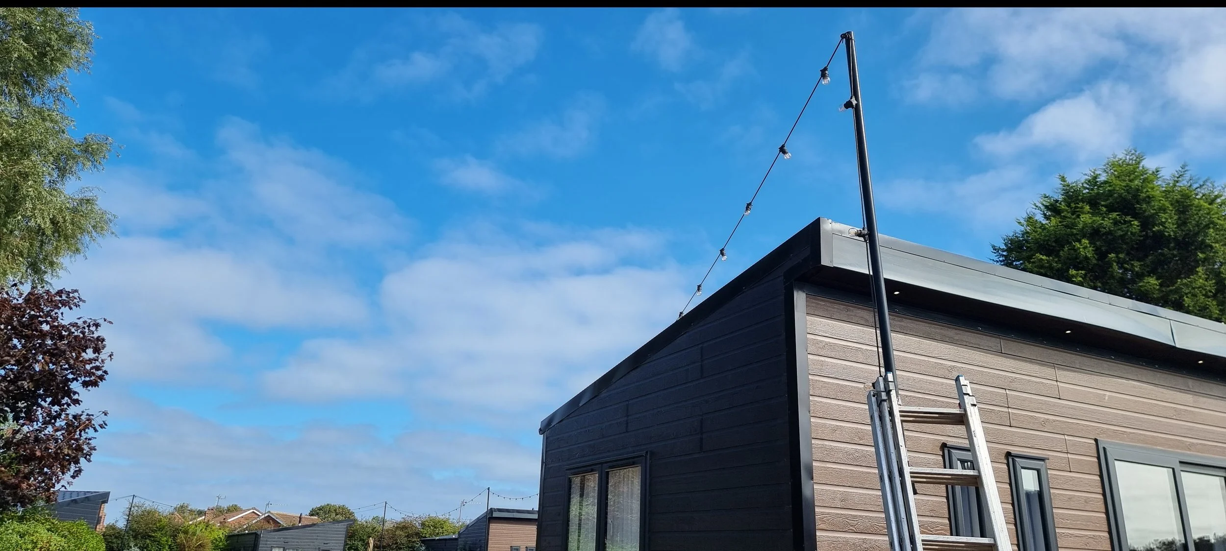A modern building with a ladder leaning against the side, an antenna pole on the roof with string lights, trees, and a blue sky with clouds.