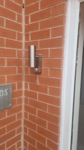 A rectangular gray doorbell with a rounded button mounted on a brick wall near a doorway.