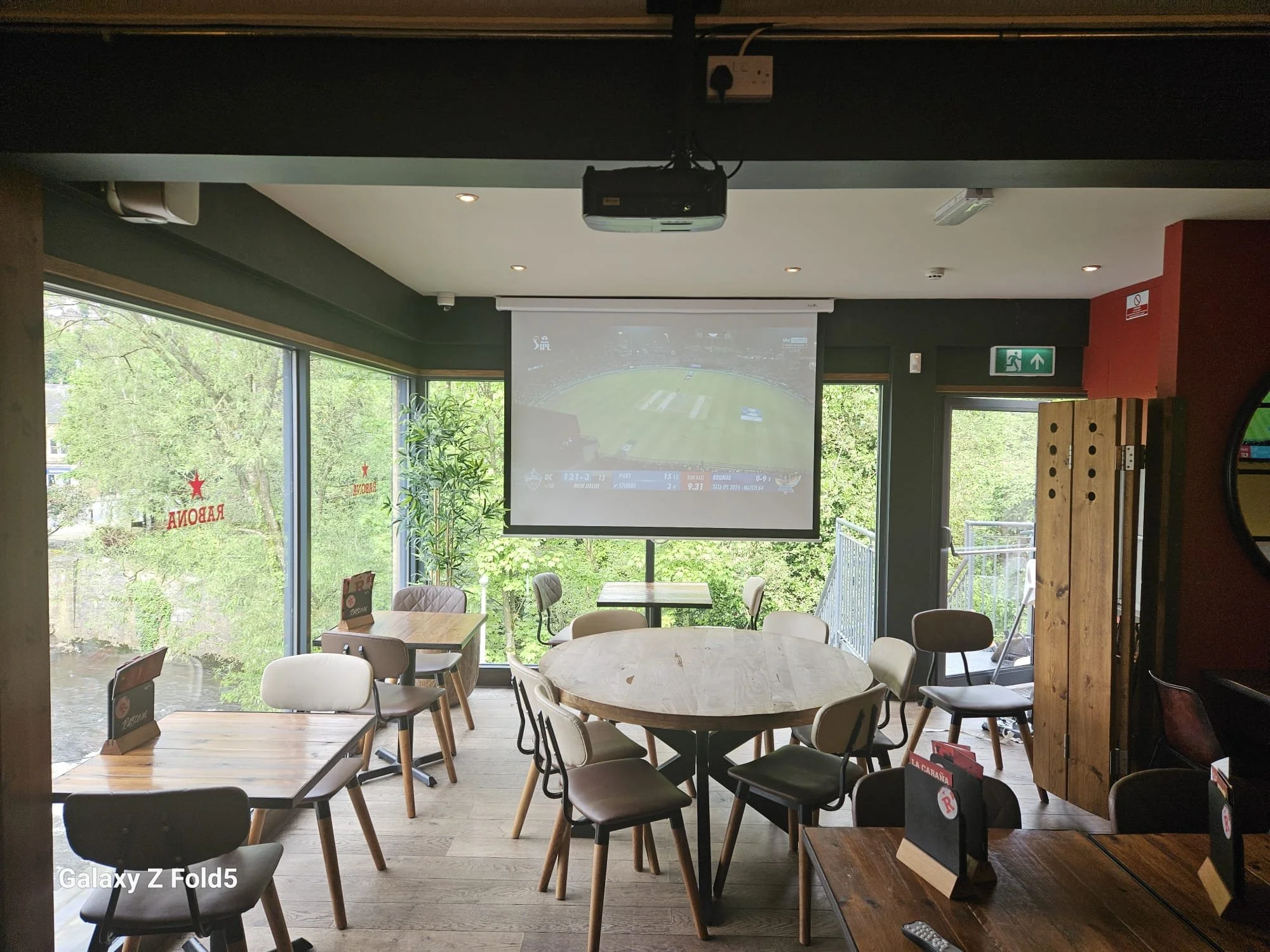 Interior of a modern cafe or restaurant with round tables and chairs, large windows showing green trees outside, and a large screen projecting a cricket match on the front wall.