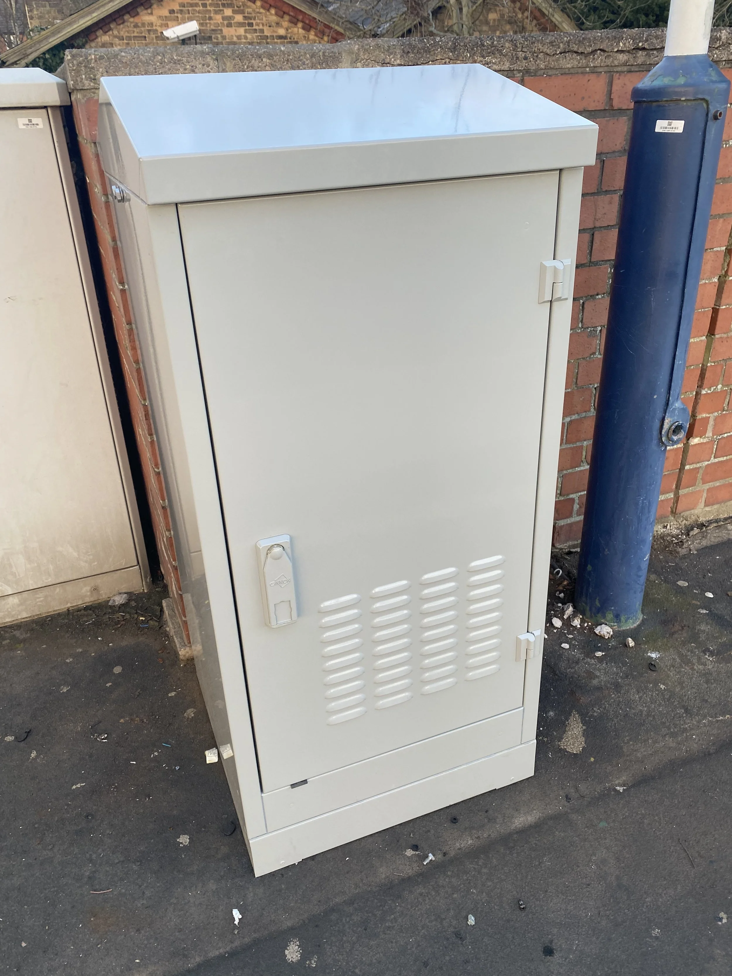 Grey electrical box or cabinet placed outdoors on pavement near a brick wall, with a blue post nearby.