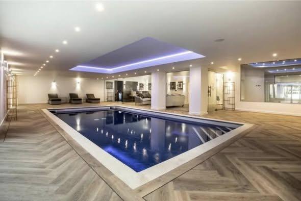 Indoor swimming pool with wooden flooring, lounge chairs, and modern lighting.