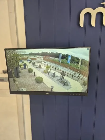 A flat-screen monitor mounted on a blue wall displaying a surveillance camera view of an outdoor park or public space with benches, bushes, and trees.