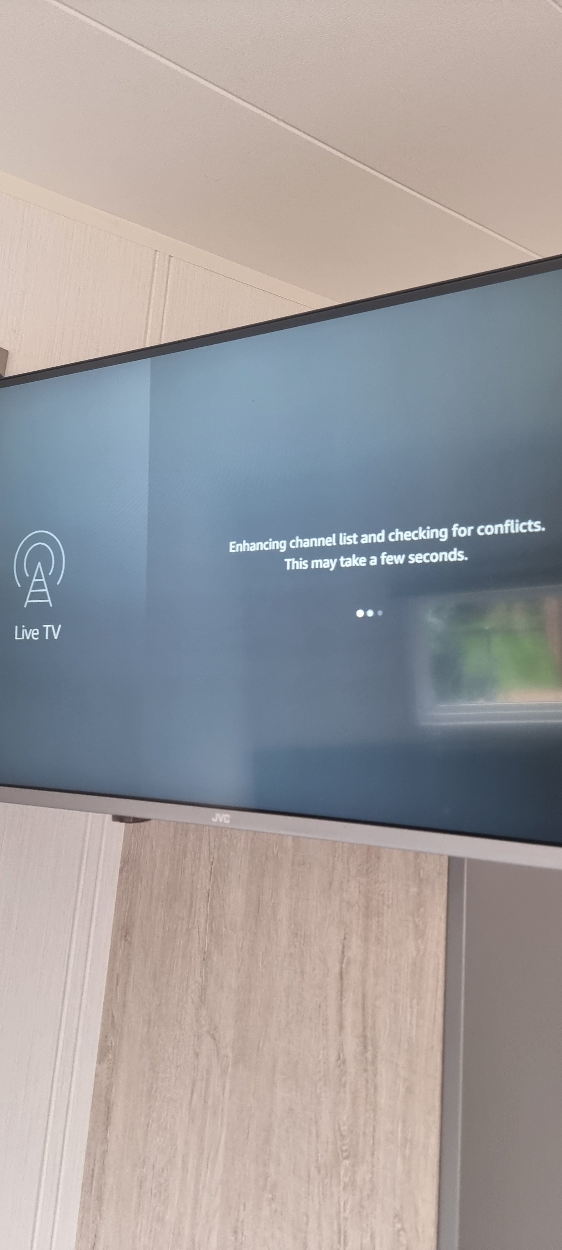 Television screen displaying a message about enhancing the channel list and checking for conflicts. The screen shows an icon for live TV and the words 'Live TV', with a partial view of a window and wall in the background.