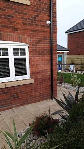 Exterior view of a brick house with a white-framed window, a black downspout, and a small garden with plants and decorative rocks.