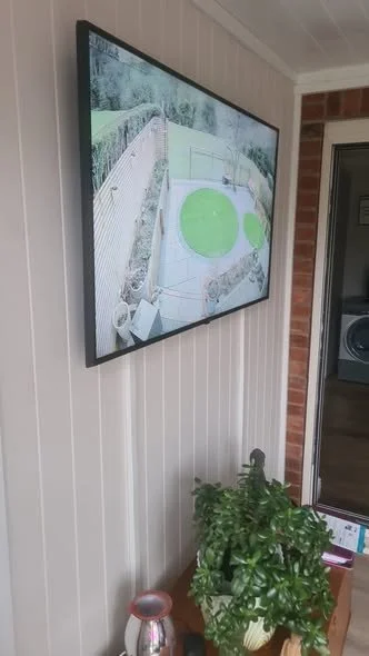 TV mounted on wall displaying a backyard with a patio, green circular area, and yard fencing. Below, a potted green plant and a ceramic vase on a wooden surface.