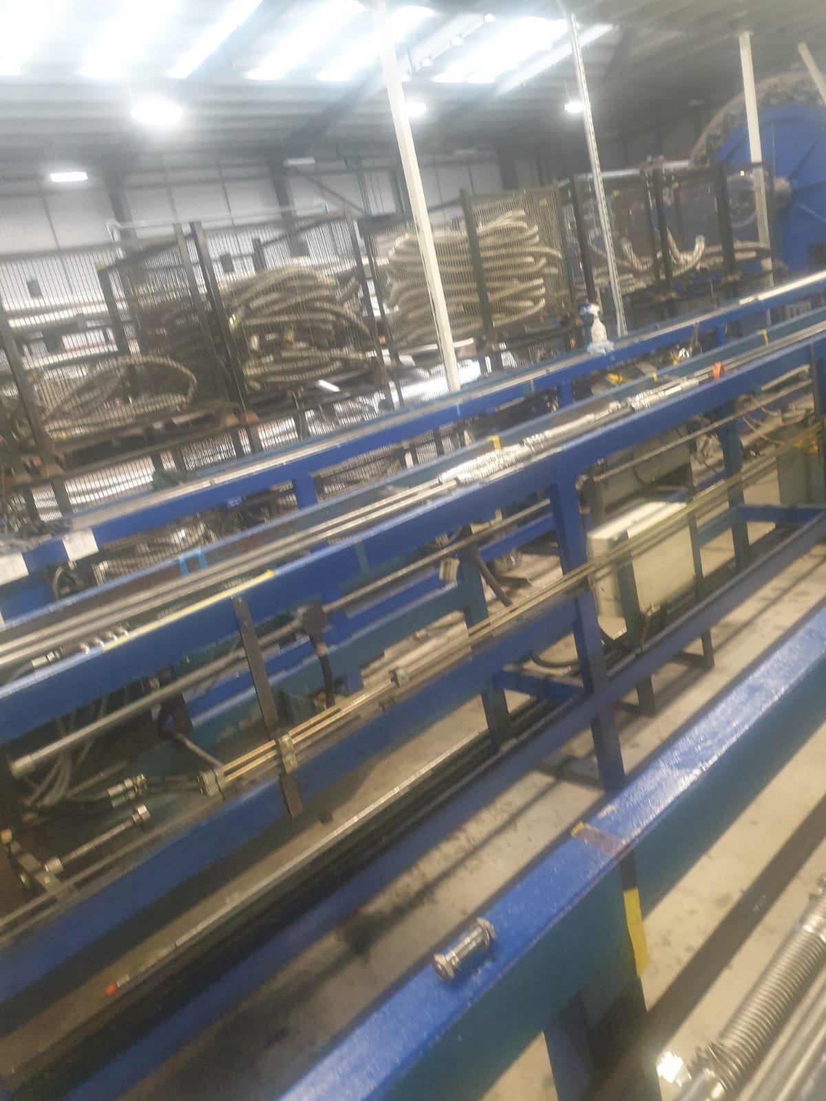 Industrial manufacturing equipment with blue metal framework and coiled metal tubes in a warehouse setting.