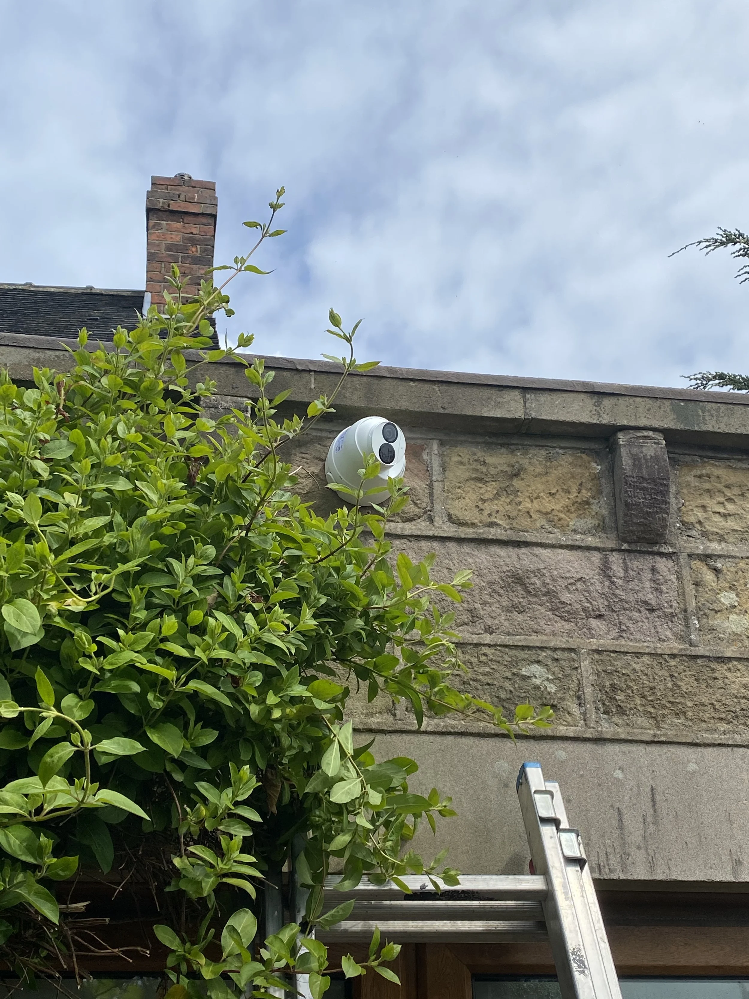 A white security camera mounted on a stone wall next to green foliage, with a ladder leaning against the wall and a chimney on the roof in the background.